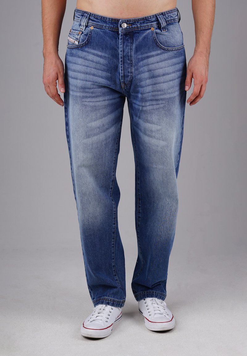 Denim jeans in medium blue with faded sections, featuring five pockets, straight leg design, and metal button closure. White sneakers visible.