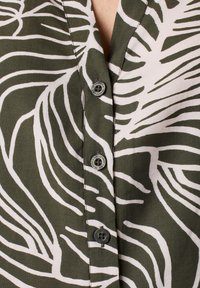 Green and white patterned blouse with a V-neckline, featuring a botanical print, and black buttons along the placket. Soft fabric texture.