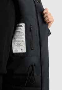 Černá péřová bunda má uvnitř zipovou kapsu, štítek s pokyny k údržbě a šňůrku na spodním lemu. Hladký materiál a funkční design.