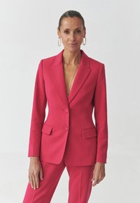 Fuchsiový oblek s padnoucím blejzrem a ladícími kalhotami. Blejzr má klopové límce, dva knoflíky a přední kapsy.