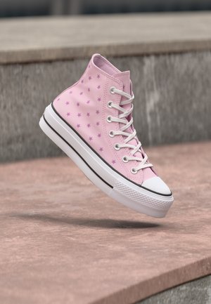 Baskets montantes roses avec motifs d'étoiles violettes, lacets blancs et semelle blanche épaisse flottant au-dessus de marches en pierre.