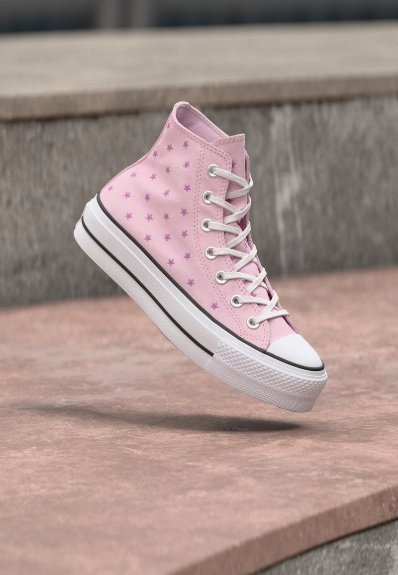 Ροζ ψηλοτάκουνο αθλητικό παπούτσι με μοτίβα από μωβ αστέρια, λευκά κορδόνια και πα χύ λευκό σόλα να αιωρείται πάνω από πέτρινα σκαλοπάτια.