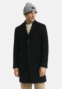 Schwarzer Wollmantel mit Notch-Revers, vier Knöpfen und Seitentaschen, getragen über einem cremefarbenen Strickpullover und kombiniert mit einer grauen Mütze.