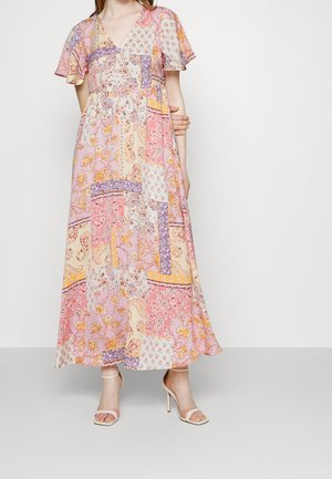 Woman wearing a patchwork floral midi dress in pink, purple, and yellow tones with flutter sleeves and white heeled sandals.