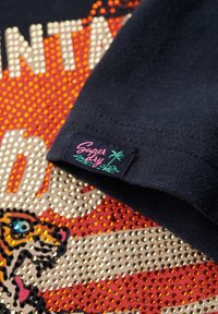 T-shirt en coton marine arborant un graphique vibrant en forme de perles représentant un tigre, accompagné d'un texte. Comprend une étiquette de marque rose avec un motif de palmier.