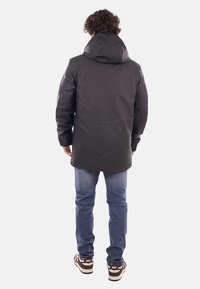 Giacca nera impermeabile con cappuccio, maniche lunghe e taglio dritto, abbinata a jeans blu e scarpe da ginnastica marroni. Design semplice, texture liscia.