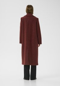 Lungo cappotto rosso realizzato in materiale testurizzato, con un ampio colletto, un'apertura centrale sul retro e maniche lunghe, abbinato a pantaloni neri.