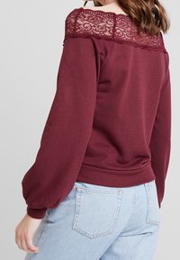 Sudadera burdeos con encaje en los hombros, mangas largas abullonadas y dobladillo acanalado. Llevada con jeans azul claro que muestran los bolsillos traseros.