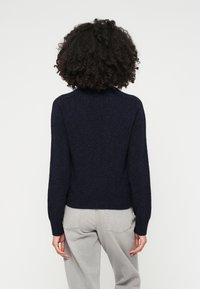 Mornarsko modra pletena pulover z visokim ovratnikom, rebrastimi manšeti in robom. Oblikovan z svetlo sivo denim hlačami, kar prikazuje sproščen kroj in mehak material.