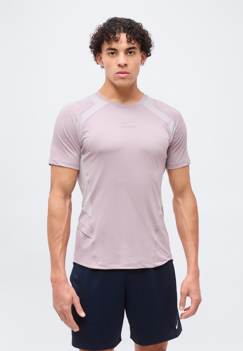 Jeune homme aux cheveux bouclés portant un t-shirt de sport violet clair et un short bleu foncé, debout devant un fond blanc uni.