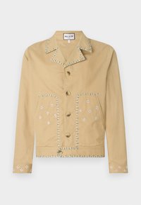 Giacca di cotone beige con colletto a revers, cinque bottoni e motivi floreali ricamati in bianco e blu su tasche e maniche.