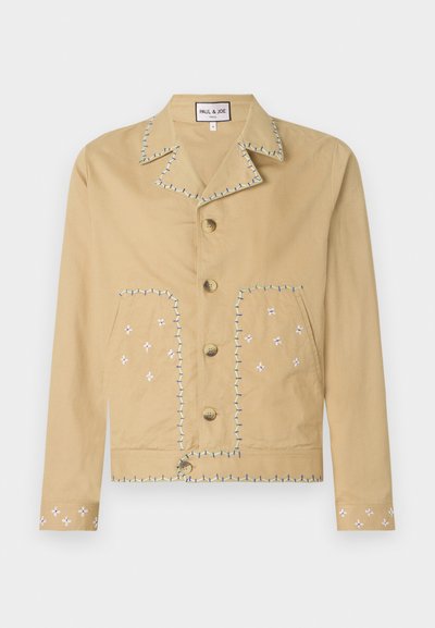 Tan cotton jacket with a notched collar, five buttons, and embroidered floral patterns in white and blue on pockets and sleeves.