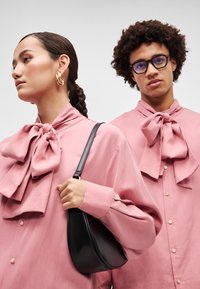 Blusas rosas com grandes laços e fechos de botão, acompanhadas de uma bolsa preta. Textura de tecido suave, usadas por duas modelos com penteados variados.