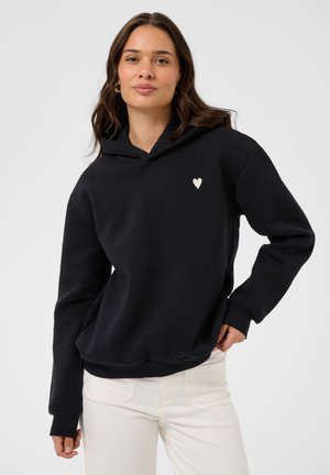 Femme aux longs cheveux bruns porte un sweat à capuche noir avec un petit cœur blanc sur la poitrine et un pantalon blanc, debout devant un fond uni.