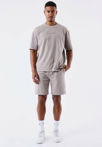Camiseta de algodón gris claro con un logo texturizado centrado en el pecho, combinada con pantalones cortos a juego. Los zapatos son blancos, junto con calcetines de longitud media.