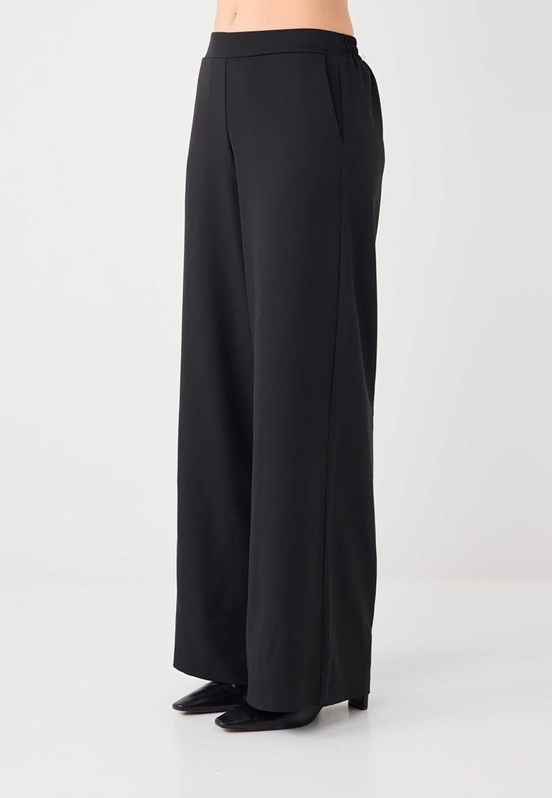 Pantalon noir à jambes larges porté par une personne avec un haut court, doté de poches latérales et associé à des chaussures noires pointues.