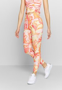 Kobieta w kolorowych legginsach w liście i dopasowanym crop topie, w białych sneakersach, stojąca na tle jednolitym.