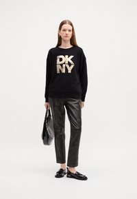 Svart sweatshirt med gull "DKNY" logo, svarte faux skinnbukser og svarte loafers. Holder en svart veske med nagler. Enkel, moderne stil.