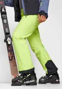 Pantalons de ski vert lime avec un accent noir aux poignets, fabriqués en matériau léger. Un ski et des bottes noires sont également visibles à côté.