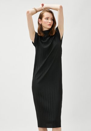 Donna che indossa un abito midi nero plissettato senza maniche, con le braccia sollevate sopra la testa, guarda di lato, su uno sfondo grigio chiaro uniforme.