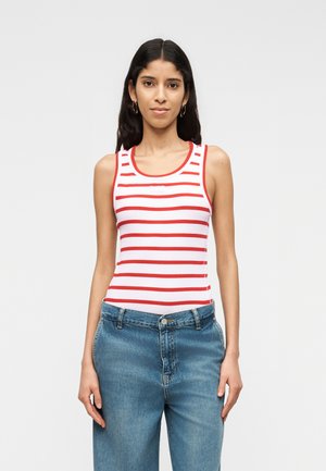 Žena stojaca, oblečená v bielom tielku bez rukávov s červenými horizontálnymi pruhmi a modrých džínsach, na jednoduchom svetlom pozadí.