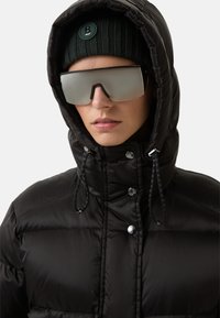 Schwarze Daunenjacke mit Kapuze, aus glänzendem Material, mit Druckknöpfen und verstellbaren Kordeln. Trägt eine dunkle Strickmütze und reflektierende Sonnenbrille.
