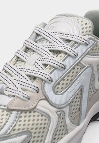 Λευκό και μπεζ αθλητικό παπούτσι από δίχτυ με επενδυμένη γλώσσα, λευκά κορδόνια με σχέδιο και ανάγλυφη λεπτομέρεια λογότυπου στην πλευρική επιφάνεια.