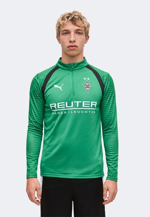 Junger Mann trägt ein grünes Puma-Sportoberteil mit langen Ärmeln, Borussia Mönchengladbach-Logo und REUTER-Sponsor-Text, steht vor einem einfarbigen Hintergrund.