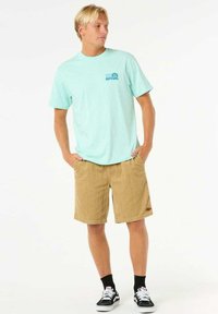 Camiseta de algodón de color azul claro con un logo azul, combinada con pantalones cortos de pana color marrón. El modelo lleva calcetas negras y zapatillas negras. Diseño sencillo.
