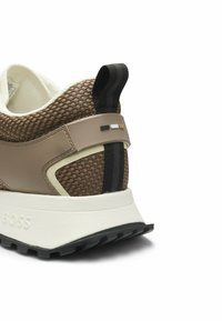 Ruskea verkko ja vaaleanbeige tennarin kantapää mustalla vetonauhalla, valkoinen pohja, musta pito ja pieniä koristeellisia mustia, valkoisia ja ruskeita neliöyksityiskohtia.