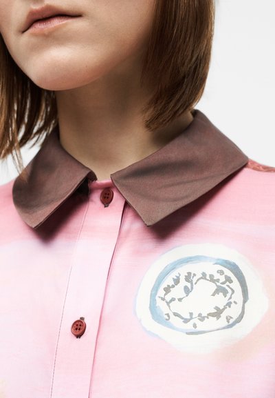 Rožinė marškinėliai su rudu apykakliu, priekiniais mygtukais ir apvaliu lapų piešiniu centre, pagaminti iš lygaus audinio.