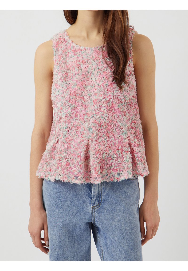 Haut péplum texturé rose sans manches avec des détails floraux multicolores porté avec un jean en denim bleu clair sur une personne debout.