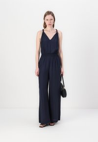 Marineblauer Jumpsuit mit weitem Bein, V-Ausschnitt und geraffter Taille. Verfügt über dünne Träger und einen glatten Stoff. Kombiniert mit einer schwarzen Handtasche.