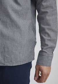 Stripet langermet skjorte i grått med hvite vertikale striper. Har en rund kant og knapp mansjett, laget av et mykt stoff.
