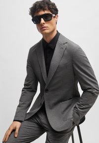 Grau strukturiertes Anzug mit passender Hose, ausgestattet mit einem reißverschlussartigen Revers und Fronttaschen. Schwarzes Hemd darunter; das Modell trägt eine dunkle Sonnenbrille.