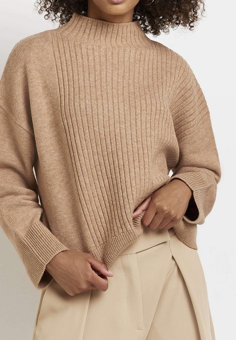 Femme portant un pull en maille côtelée beige associé à un pantalon plissé beige clair, ajustant l'ourlet du pull.