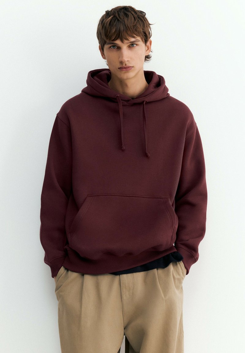 Jeune homme aux cheveux bruns portant un sweat à capuche bordeaux et un pantalon beige, debout les mains dans les poches sur un fond blanc uni.