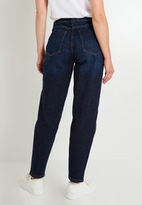 Pantalones vaqueros de mezclilla de cintura alta en azul oscuro con corte recto, que cuentan con dos bolsillos traseros y un bajo limpio. Combinados con zapatillas blancas.