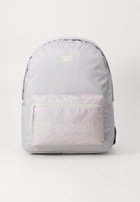 Mochila cinza-claro com uma textura suave, apresentando um bolso frontal com fecho em rosa e o logótipo "Reebok" em branco, além de alças ajustáveis em preto.