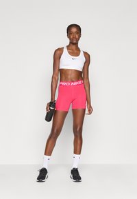 Sportinė apranga, kurią sudaro balta sportinė liemenėlė ir ryškiai rožiniai šortai su plačiu juosmeniu, pažymėtu "PRO NIKE". Taip pat yra juodas vandens butelis.