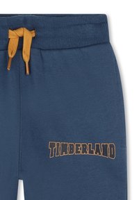 Pantalon de survêtement bleu avec une ceinture côtelée et un cordon de serrage orange. Logo noir "TIMBERLAND" imprimé sur le côté gauche près du bas.