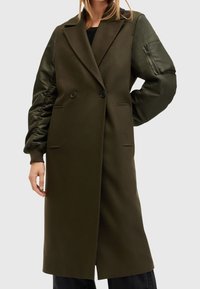 Abrigo verde oliva con diseño estructurado, que presenta un frente de doble botonadura, bolsillos laterales y mangas abullonadas con textura satinada.