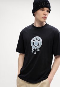 Svart bomull t-skjorte med et rundt grafisk motiv som har et dryppende smilefjes og "HGO" tekst i lys blått og svart. Casual design.