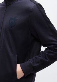 Σκούρο ναυτικό μπλε ζακέτα με φερμουάρ, κατασκευασμένη από λείο ύφασμα, με υφασμένο έμβλημα στο στήθος, ριμπ μανσέτες και πλαϊνές τσέπες.