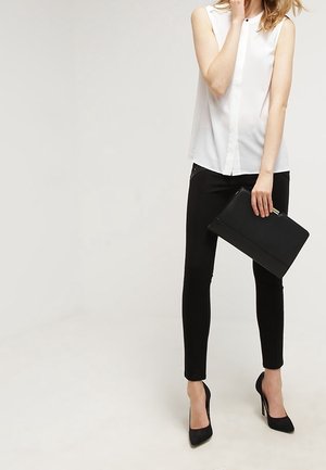 Femme portant un chemisier blanc sans manches, un pantalon noir slim et des talons hauts noirs, tenant une pochette noire dans sa main droite.