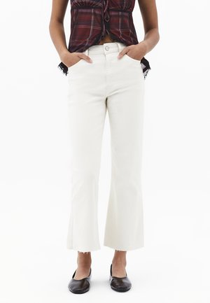 Osoba nosząca białe dżinsy o kroju flare do połowy łydki, związany bordowy kraciasty top bez rękawów, czarne baleriny, ręce w kieszeniach.