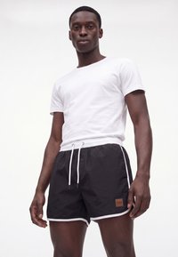 Mann trägt ein schlichtes weißes Kurzarm-T-Shirt und schwarze Shorts mit Kordelzug und weißem Rand, steht vor einem einfarbigen hellen Hintergrund.