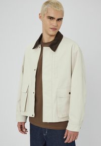 Veste de couleur crème avec un col en velours côtelé brun, fermeture à pression à l'avant, deux poches latérales et des détails de couture visibles. Portée sur une chemise marron.