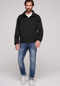 Giacca nera con zip e colletto alto, due tasche laterali, abbinata a jeans chiari e sneaker bianche, con una vestibilità comoda.