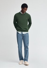 Grön stickad tröja med V-hals, kombinerad med en ljusblå skjorta med knappar, blå jeans och beige sneakers. Modellen står och ler.